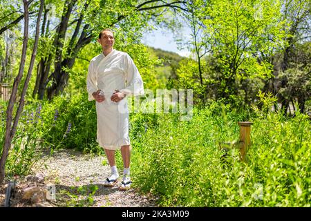 Jeune homme heureux en robe blanche yukata spa costume ou robe debout dans le jardin extérieur au Japon avec vue sur la nature Banque D'Images