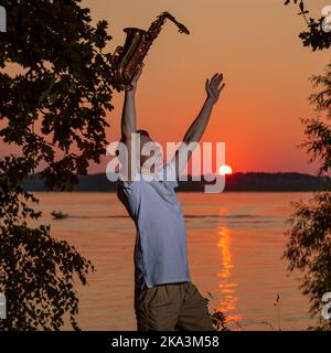 Un jeune homme avec un saxophone a levé les mains, profitant du coucher du soleil. Banque D'Images