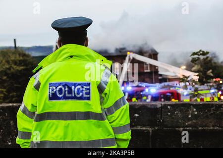 Un policier portant une veste haute visibilité regarde une brigade de pompiers s'attaquer à un incendie dans un bâtiment de l'ancienne école Banque D'Images