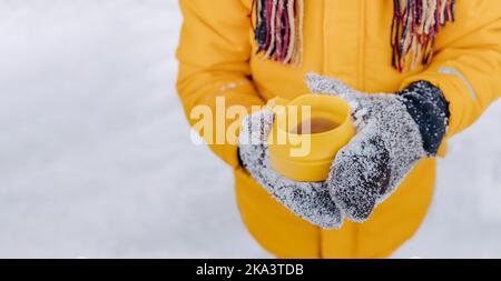 Gros plan des mains tenant une tasse de thé chaud jaune sur fond de neige. Garçon en gants de neige tricotés et écharpe avec mug chaud. Thème des vacances d'hiver avec Banque D'Images