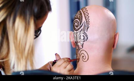 Une fille, maître de tatouage, l'artiste mehendi fait le dessin de henné tatouage sur le cuir chevelu de l'homme blanc, épaule, cou.la solution de henné sèche sur la peau. Photo de haute qualité Banque D'Images