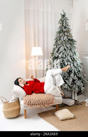 Une femme fait ses achats de Noël en ligne. Elle est assise sur le canapé à la maison, parlant au service de livraison, arbre de Noël en arrière-plan. Banque D'Images