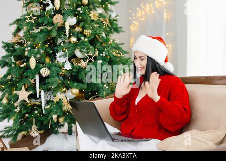 Une jeune femme heureuse dans un chapeau de père Noël, parlant à ses amis lors d'un appel vidéo virtuel et leur signe, célèbre le nouvel an lors d'une fête en vacances Banque D'Images