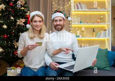 Portrait de famille heureux acheteurs en ligne, couple amoureux à la maison assis sur le canapé près de l'arbre pour les vacances de Noël et le nouvel an, regardant l'appareil photo et souriant, tenant l'ordinateur portable et la carte de crédit bancaire. Banque D'Images