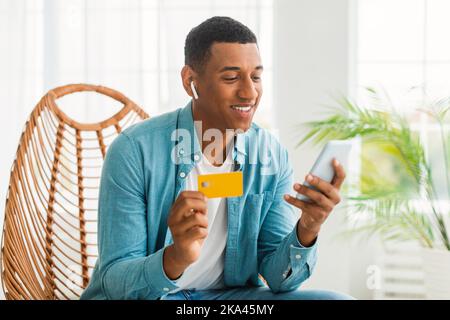 Un jeune homme afro-américain heureux avec casque sans fil utilise un smartphone et une carte de crédit Banque D'Images