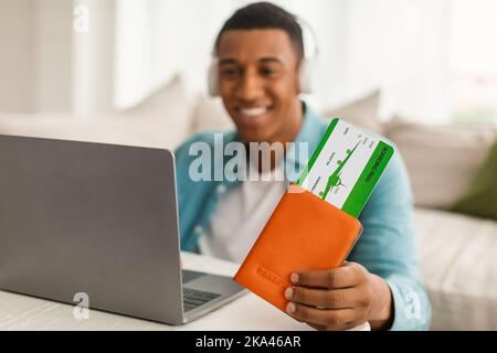 Un jeune afro-américain heureux, dans un casque sans fil, montre des passeports et des billets d'avion, regarde l'ordinateur Banque D'Images