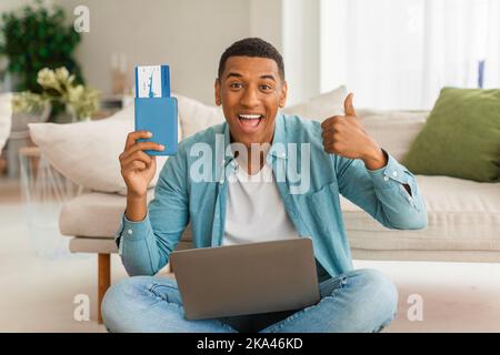 Joyeux homme afro-américain millénaire avec bouche ouverte montre passeport et billet d'avion utilise ordinateur portable Banque D'Images