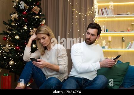 Un couple de familles se querellé à Noël assis à la maison sur un canapé, mari et femme ne parlant pas les uns avec les autres, tenant des téléphones dans les mains parcourant les réseaux sociaux, près de l'arbre de Noël. Banque D'Images