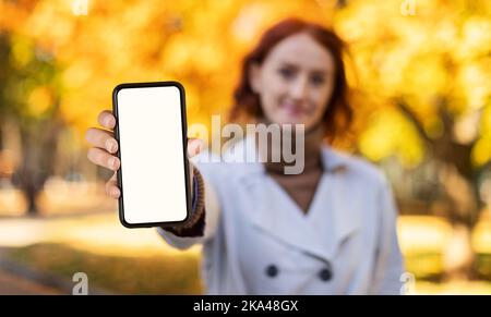 Jeune femme européenne souriante aux cheveux rouges en imperméable montre un smartphone avec écran vierge dans le parc de la ville Banque D'Images