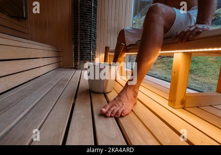 Relaxant homme enveloppé serviette blanche assis et transpirant sur le banc en bois dans le sauna finlandais chaud avec des pierres chaudes et appréciant le soin agréable du corps tempera Banque D'Images