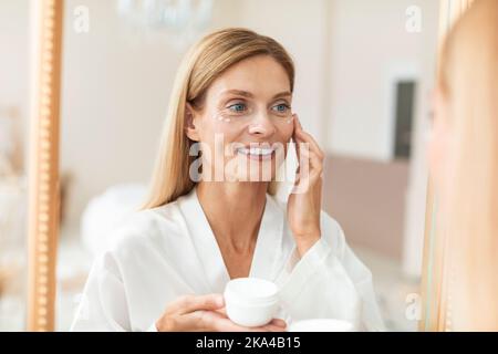 Bonne blonde moyenne femme en peignoir avec crème hydratante pour les yeux, regardant le miroir, appréciant les soins du visage routine Banque D'Images