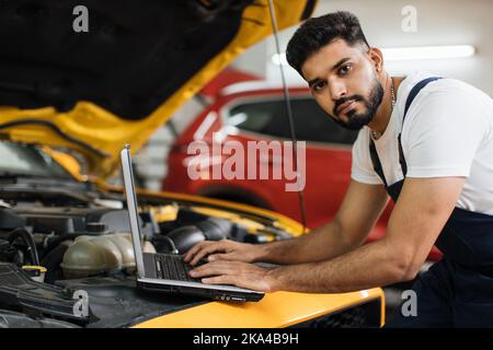 Jeune homme mécanicien de voiture entretient le véhicule en utilisant l'ordinateur portable de diagnostic pour vérifier les problèmes de moteur, la technologie moderne dans l'atelier de réparation de voiture. Banque D'Images