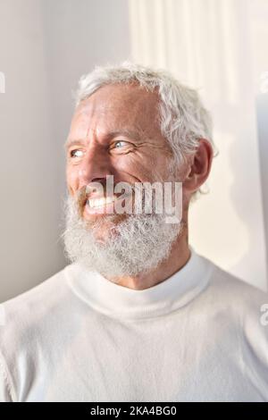 Homme âgé souriant, barbu, debout sur un mur blanc en plein soleil et regardant la fenêtre. Banque D'Images