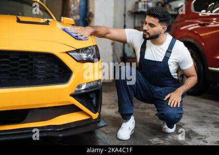 Homme barbu de lavage de voiture, portant des vêtements de protection uniforme combinaisons, nettoyage moderne jaune voiture de luxe avec un tissu microfibre. Habillage de voiture ou concept de véhicule. Banque D'Images