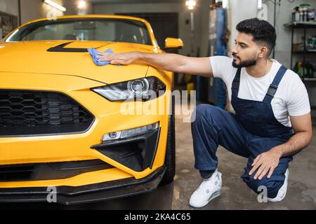 Homme barbu de lavage de voiture, portant des vêtements de protection uniforme combinaisons, nettoyage moderne jaune voiture de luxe avec un tissu microfibre. Habillage de voiture ou concept de véhicule. Banque D'Images