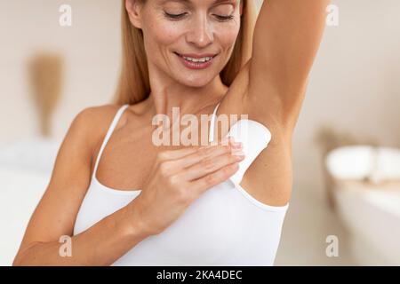 Belle femme d'âge moyen appliquant un bâton antisudorifique sur l'aisselle pour la protection contre la transpiration, debout dans la chambre à coucher Banque D'Images