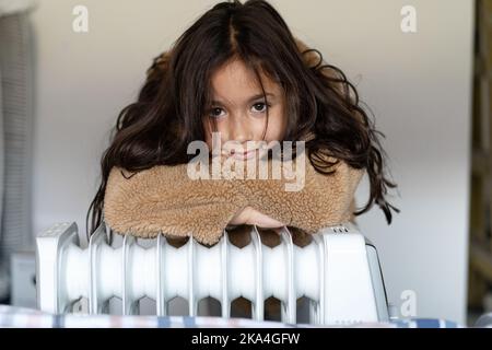 Enfant vêtu d'un manteau en fausse fourrure beige chaud près d'un chauffe-maison en hiver froid. Utilisation du chauffage à la maison en hiver. Belle petite fille chauffe près du chauffage et regarde dans l'appareil photo. Banque D'Images