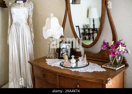 Commode et miroir anciens avec un assortiment de bouteilles de parfum, photo de mariage, fleurs, lampe, et une robe de mariage dans le coin de la chambre d'une mariée. Banque D'Images