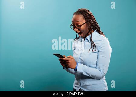 Joyeux africain américain avec un smartphone surpris par les e-mails reçus. Jeune femme souriante et joyeuse avec un téléphone portable moderne, stupéfaite par les médias sociaux après la portée. Banque D'Images