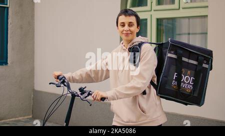 Happy Woman Food Courier assis à la porte d'entrée pour livrer la commande de repas à emporter, portant sac à dos avec le déjeuner. Employé du service de livraison livrant des aliments fastfood dans un sac thermique et à vélo. Banque D'Images