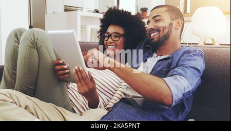 Naviguer sur Internet ensemble. Un jeune couple joyeux assis sur un canapé tout en naviguant sur une tablette numérique dans le salon. Banque D'Images