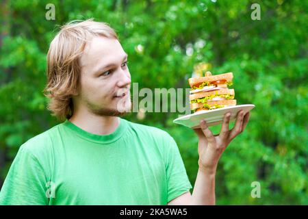 Jeune homme tenant un gros sandwich à la saucisse sur l'assiette. L'étudiant mange de la nourriture rapide dans un paquet sur fond de feuillage vert. Aliments indésirables rapides. Un homme très affamé. Concept de régime. Banque D'Images