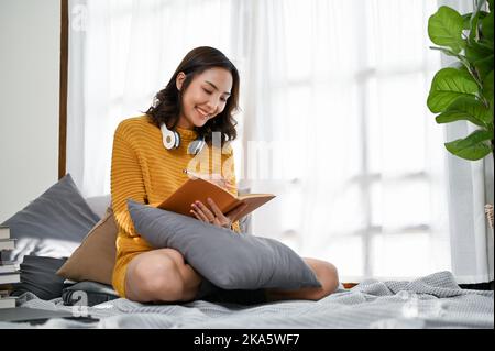 Jeune femme asiatique attrayante et gaie vêtue de vêtements décontractés, écrivant son journal ou prenant des notes sur son carnet tout en se relaxant dans le salon. Banque D'Images