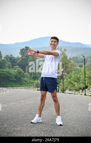 Beau et heureux jeune asiatique dans les vêtements de sport étirant ses bras et son corps, se réchauffant avant l'exercice ou se refroidissant après la course. Sport de plein air Banque D'Images