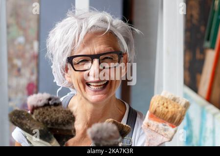 souriante heureuse femme portrait, fière artiste, dans ses années 50 avec cheveux gris et lunettes noires et beaucoup de pinceaux, espace de copie Banque D'Images