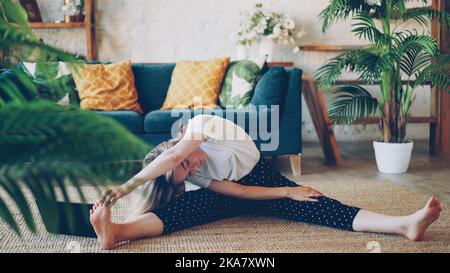 Flexible jeune blond fait des exercices d'étirement à la maison assis sur le sol dans l'appartement moderne. La fille se plie de la tête au genou en tenant les orteils et en s'étirant vers l'arrière et les jambes. Banque D'Images