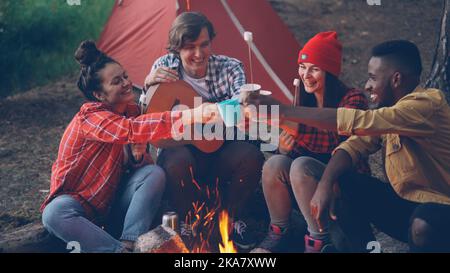 Joyeux jeunes amis sont de clinking verres avec des boissons assis autour du feu tente nera dans la forêt avec guimauve chaude sur des bâtons, homme souriant tient la guitare. Banque D'Images