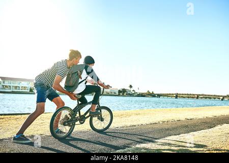 Parfois, vous avez besoin d'une poussée de votre frère aîné. Prise de vue en longueur d'un jeune garçon expliquant à son frère cadet comment faire un vélo le long d'un lagon. Banque D'Images