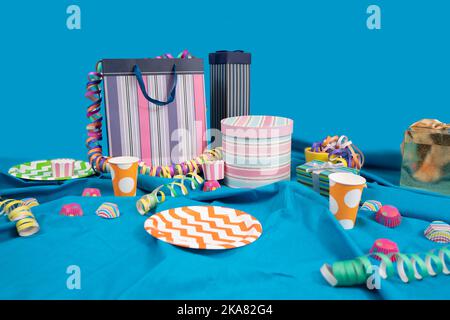 Table de fête désordonnée avec sacs et boîtes cadeaux, banderoles, assiettes et tasses en plastique sur une table avec une nappe bleue sur un fond bleu clair Banque D'Images