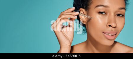 Femme africaine tenant un compte-gouttes de pipette avec de l'huile essentielle ou du sérum près de son visage pour hydrater et hydrater la peau Banque D'Images