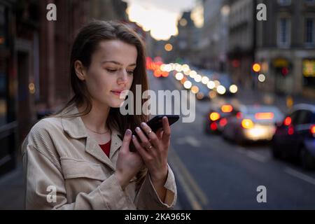 Une jeune femme se tenant dans une rue de la ville à Newcastle upon Tyne dans la soirée tout en parlant à un ami sur son téléphone portable. Elle attend un taxi. Banque D'Images