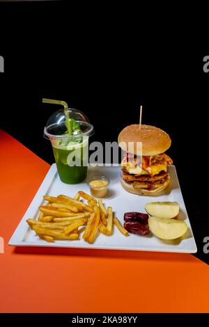 Un gros plan d'un hamburger, de frites et d'une sauce avec des morceaux de pommes et des dattes avec une tasse de jus de menthe sur un plateau blanc Banque D'Images