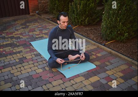 Vue de dessus d'un athlète caucasien, un yogi assis pieds nus dans lotus pose sur un tapis de yoga, médite avec le rosaire Banque D'Images