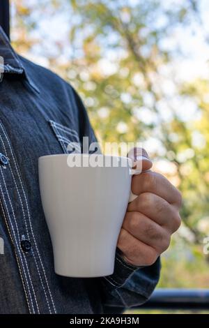 main de l'homme tenant une tasse de café à côté d'une fenêtre avec arrière-plan d'automne dehors Banque D'Images