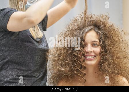 Le maître de cheveux dans le masque de visage fait une coiffure bouclés pour une femme souriante. Portrait d'une belle fille caucasienne avec des cheveux bouclés coulant. Le concept de salon de beauté. Photo de haute qualité Banque D'Images