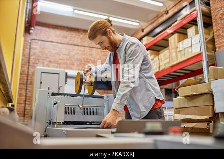 Vue latérale d'un jeune homme qui a mis en place une machine à imprimer à rouleaux dans un atelier industriel, espace de copie Banque D'Images