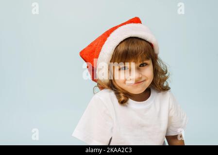 Drôle mignon fille dans un chapeau de père noël se tient sur un fond bleu avec l'espace de copie. Concept de noël et nouvel an Banque D'Images