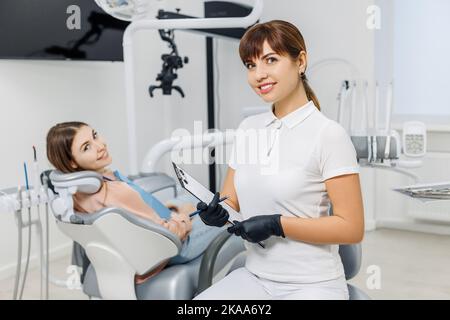 Dentiste amical avec un comprimé sur l'arrière-plan du patient pendant l'examen des dents. Clinique dentaire moderne. Concept de soins buccodentaires professionnels, consultant Banque D'Images