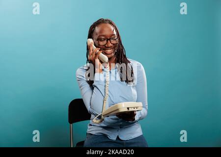 Jeune femme adulte confiante et positive répondant aux appels des clients pour des services de conseil. Un opérateur de téléphone fixe souriant ayant une conversation sur un téléphone filaire sur fond bleu. Banque D'Images