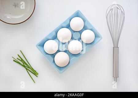 Hachez des œufs géants blancs frais dans un carton d'œufs en céramique bleue avec fouet, ciboulette et bol en composition de ponte plate. Ingrédients sains sur fond blanc Banque D'Images