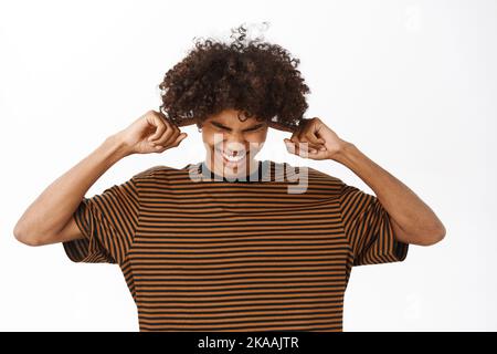 Un jeune homme agacé ferme ses oreilles avec les doigts, les bouchons d'oreilles et les grimaces du bruit fort, se tient sur fond blanc Banque D'Images