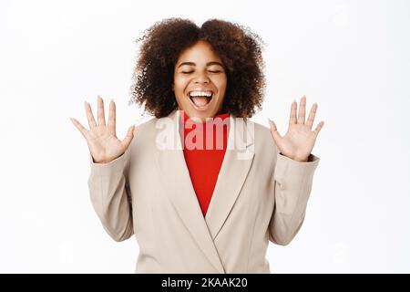 Une femme noire excitée crie de joie, porte un costume, célèbre et triomphe, se dresse sur fond blanc Banque D'Images