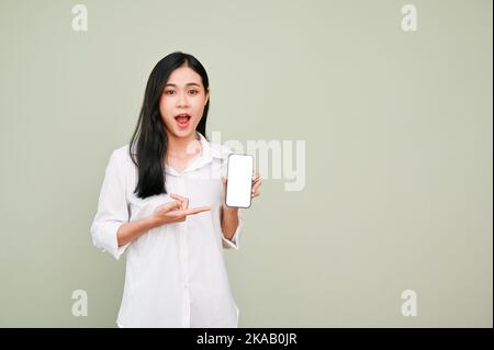 Émerveillée, choquée et surprise, la jeune femme asiatique en chemise blanche pointe le doigt vers son smartphone, écran blanc, isolée sur le dos gris Banque D'Images