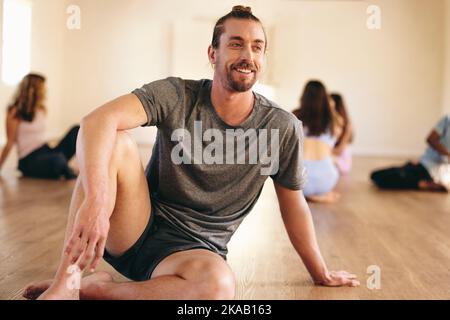 Un jeune homme heureux assis dans un studio de yoga avec un groupe de personnes en arrière-plan. Jeune homme souriant ayant une séance de yoga avec sa classe dans un communii Banque D'Images