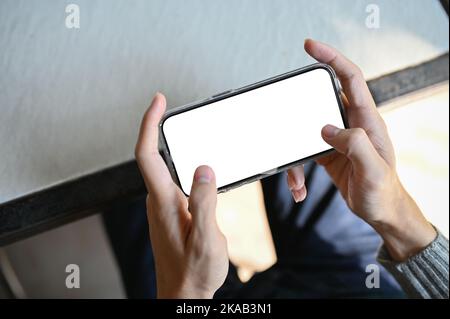 Les mains d'un homme se touchent sur l'écran du téléphone, jouent à un jeu ou bavardent avec ses amis. La maquette de l'écran blanc du smartphone en position horizontale est mal Banque D'Images