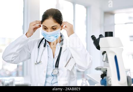 Il est essentiel pour les travailleurs de laboratoire de porter les vêtements de protection appropriés. Un jeune scientifique mettant un masque chirurgical tout en travaillant dans un laboratoire. Banque D'Images